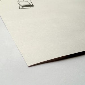 May include: A close-up of a corner of a piece of paper with a sketch of a chair in the upper right corner. The paper is off-white and has a slight texture. The edge of the paper is dark.