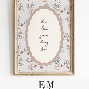 May include: A framed print with a light floral pattern background and an oval shape in the center. The oval contains the handwritten text "slow down you're doing fine". The frame is a light brown color. The bottom of the print has the text "EM Atelier".