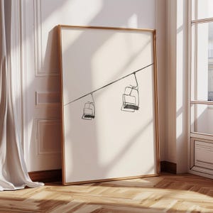 May include: A framed art print with a minimalist line drawing of two ski lift chairs hanging from a cable. The artwork is in black and white, with a gold frame. The print is leaning against a wall with a white curtain and a wooden floor.