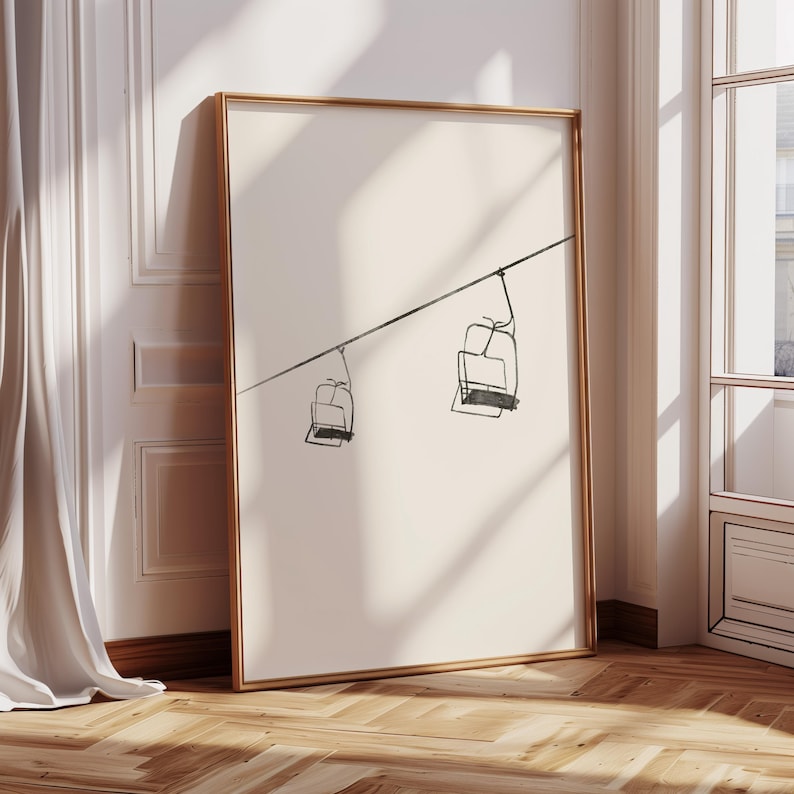 May include: A framed art print with a minimalist line drawing of two ski lift chairs hanging from a cable. The artwork is in black and white, with a gold frame. The print is leaning against a wall with a white curtain and a wooden floor.
