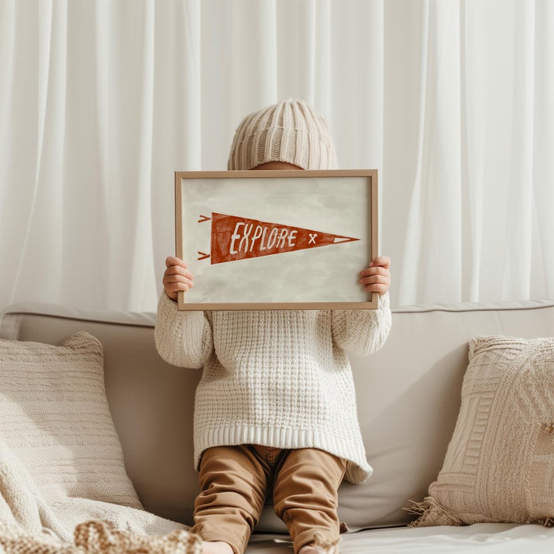 May include: A framed print with the word "EXPLORE" in a red pennant design. The print is held by a person wearing a white sweater, tan pants, and a knit cap. The background includes a sofa and curtains.
