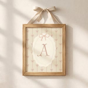 May include: A framed print with the letter "A" in pink, set against a cream background with a bow pattern. The print is in a light brown wooden frame, suspended by a matching ribbon. The overall design has a vintage feel.