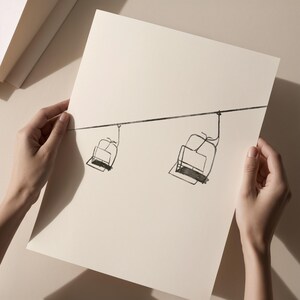 May include: A monochrome line drawing of two ski lift chairs hanging from a cable. The artwork is on cream-coloured paper. The image is held by two hands, against a neutral background.