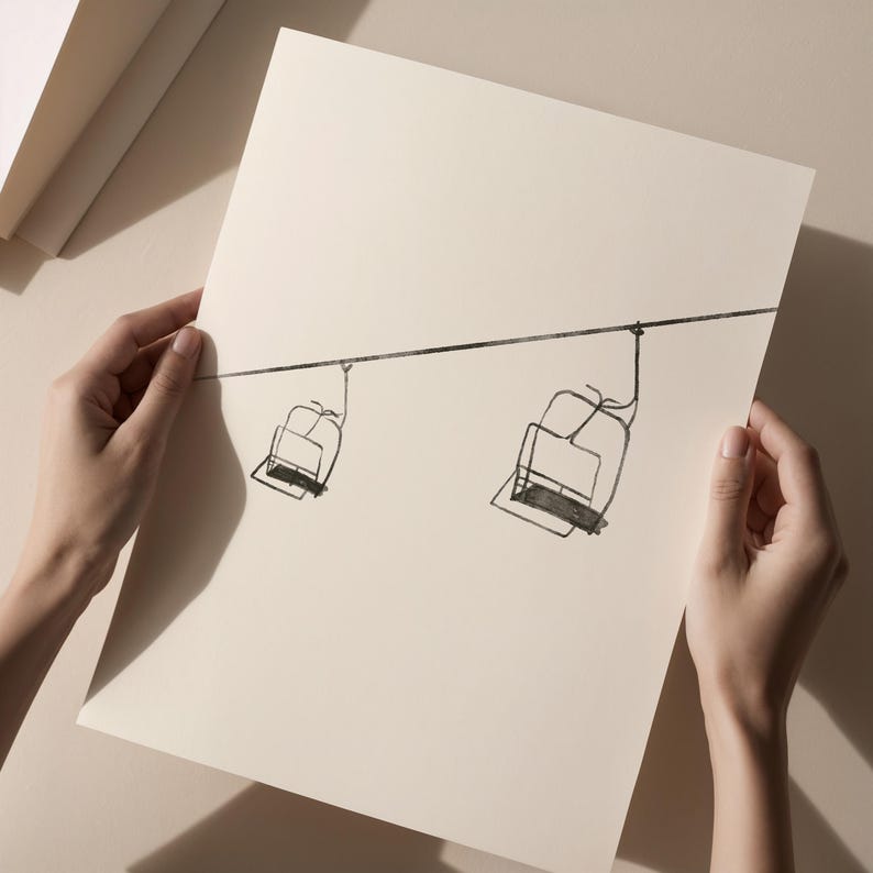 May include: A monochrome line drawing of two ski lift chairs hanging from a cable. The artwork is on cream-coloured paper. The image is held by two hands, against a neutral background.