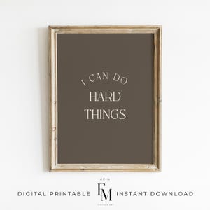 May include: Framed wall art with the text "I CAN DO HARD THINGS" in a white, elegant font. The background is a solid, muted brown. The frame is a light, distressed wood. The bottom of the frame has the text "DIGITAL PRINTABLE" and "INSTANT DOWNLOAD".