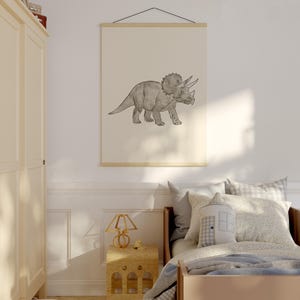 May include: A framed print of a Triceratops dinosaur in a child's bedroom. The artwork is a grayscale illustration of the dinosaur. The print is hung on a wall above a bed with pillows and a blanket. A wooden nightstand and lamp are also visible.