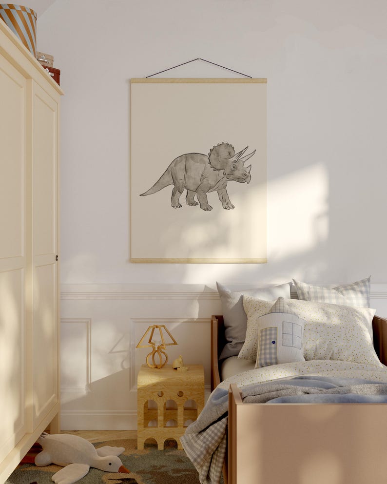 May include: A framed print of a Triceratops dinosaur in a child's bedroom. The artwork is a grayscale illustration of the dinosaur. The print is hung on a wall above a bed with pillows and a blanket. A wooden nightstand and lamp are also visible.
