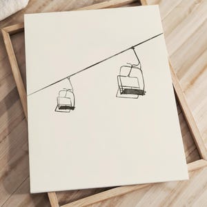May include: A framed art print with a minimalist line drawing of two ski lift chairs hanging from a cable. The artwork is in black on a cream background, set in a natural wood frame. The piece suggests winter and outdoor activities, suitable for a home or chalet.