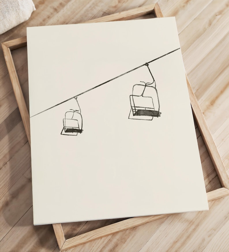 May include: A framed art print with a minimalist line drawing of two ski lift chairs hanging from a cable. The artwork is in black on a cream background, set in a natural wood frame. The piece suggests winter and outdoor activities, suitable for a home or chalet.