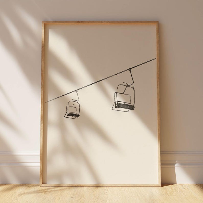 May include: Framed art print with a minimalist line drawing of two ski lift chairs on a cable. The black drawing is set against a cream background, within a light wood frame. The artwork suggests winter sports and outdoor activities, suitable for home decor.