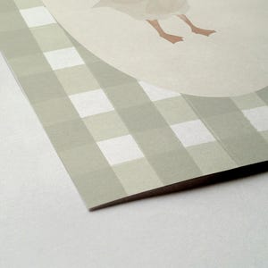 May include: A printed card with a light green and white gingham pattern. A light beige oval shape with a goose illustration is in the upper right corner. The goose's feet are visible. The card's corner is in focus.