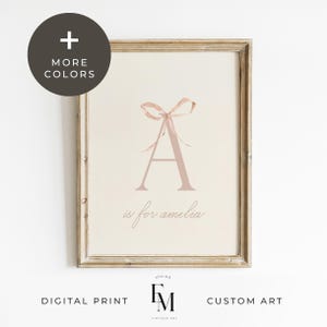 Puede incluir: Una impresión de arte de guardería imprimible con un lazo de cinta rosa y la letra A en color rosa claro. El texto "A is for Amelia" está escrito en cursiva debajo de la letra. La impresión está enmarcada en un marco de madera claro.