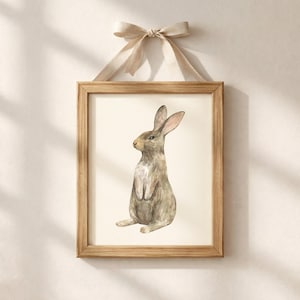 May include: A framed wall art print featuring a watercolor illustration of a brown rabbit sitting upright. The artwork is encased in a light-toned wooden frame and is suspended by a beige fabric ribbon tied in a bow at the top of the frame.