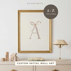 May include: A framed wall art print featuring a large letter "A" in a soft taupe color, adorned with a ribbon. The print is in a gold frame. Text on the print reads "A-Z INCLUDED" and "CUSTOM INITIAL WALL ART".