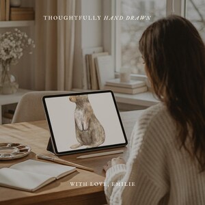 May include: A tablet screen displays a watercolor illustration of a brown rabbit in profile. The text THOUGHTFULLY HAND DRAWN and WITH LOVE, EMILIE appears on the screen. A paintbrush and an open notebook rest on the wooden table surface.