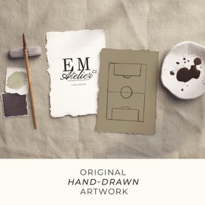 May include: An artistic composition featuring hand-drawn artwork. The image includes a soccer field sketch, a piece of paper with the text "EM Atelier co HOME PRINTS", paint swatches, a paintbrush, and a small ceramic dish with dark brown paint.