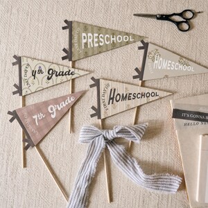 Könnte beinhalten: Eine Sammlung dreieckiger Wimpel an Holzstäben mit Aufschriften wie Preschool, 4th Grade, 7th Grade und Homeschool. Die Wimpel haben verschiedene Muster und Farben, darunter Olivgrün, Creme und Altrosa, ergänzt durch eine gestreifte Stoffschleife.