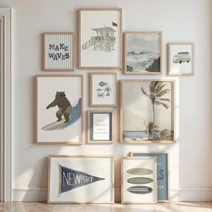 May include: A gallery wall arrangement of eleven framed prints featuring coastal and nautical themes, including a bear surfing, a lifeguard tower, palm trees, surfboards, a Newport pennant, and text-based art, all in light wood frames.