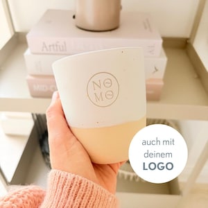 May include: A two-toned ceramic cup with a matte white upper section and a light beige lower section. The front features a circular logo with the text NOME inside. A white circular overlay on the right displays the text auch mit deinem LOGO.