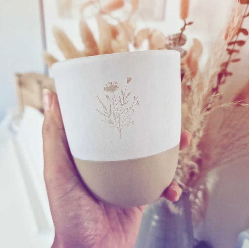 May include: A two-toned ceramic mug featuring a matte white upper section and a smooth tan lower section. The white portion is decorated with a delicate, fine-line botanical illustration of a wildflower bouquet in a soft, muted gold color.