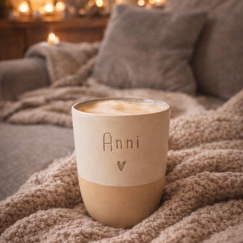 May include: A beige ceramic mug with a smooth upper section and a textured lower section. The word Anni is embossed on the front above a small heart symbol. The mug is filled with a light-colored frothy beverage, showing a simple cylindrical shape.