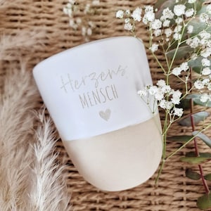 May include: A two-toned ceramic mug with a matte beige base and a white glazed upper section. The white portion features the grey text Herzens MENSCH and a small grey heart icon. The mug is positioned next to sprigs of white baby's breath flowers.