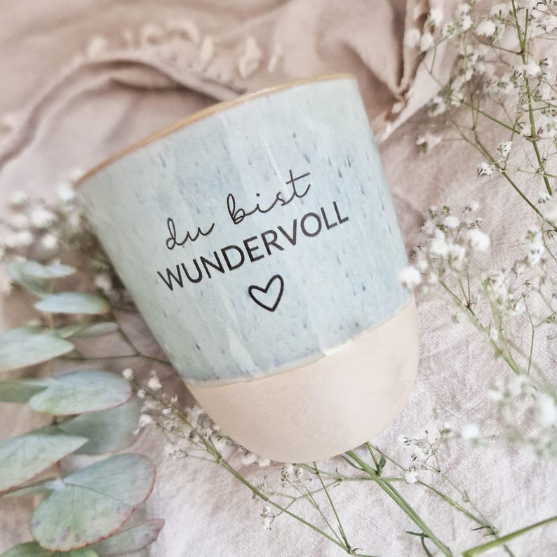May include: A ceramic mug with a speckled light blue glazed upper section and a matte beige base. The mug features the black printed text "du bist WUNDERVOLL" followed by a small heart symbol. The design is simple, cylindrical, and handleless.