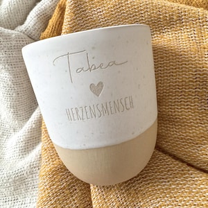 May include: A two-toned ceramic mug with a speckled white upper section and a smooth tan base. The white portion features the engraved name Tabea, a small heart symbol, and the word HERZENSMENSCH in a clean, sans-serif font centered on the front.
