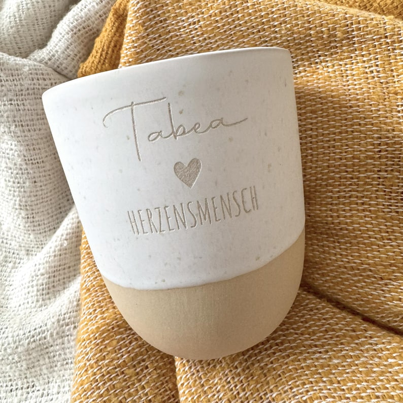 May include: A two-toned ceramic mug with a speckled white upper section and a smooth tan base. The white portion features the engraved name Tabea, a small heart symbol, and the word HERZENSMENSCH in a clean, sans-serif font centered on the front.