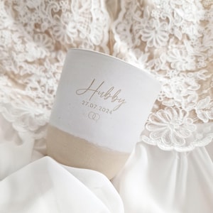 May include: A white and beige ceramic tumbler featuring the word Hubby in a script font, the date 27.07.2024, and a small icon of two interlocking rings. The cup has a smooth white upper section and a natural beige base.
