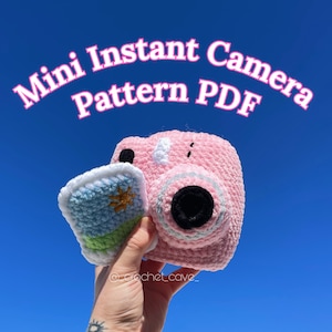 Könnte beinhalten: Eine rosa gehäkelte Mini-Sofortbildkamera mit einem blau-grünen gehäkelten Foto. Die Kamera hat ein schwarzes Objektiv und ein weißes gehäkeltes Detail oben. Der Text "Mini Instant Camera Pattern PDF" ist in rosa Buchstaben auf blauem Hintergrund geschrieben.