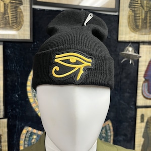 May include: Black knit beanie featuring a gold Eye of Horus patch. The hat is displayed on a white mannequin head. The background includes framed Egyptian art and a dark blue patterned fabric.