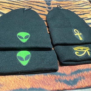 May include: Four black knit beanies with different embroidered designs. Two beanies have green alien faces, one has a gold ankh symbol, and one has a gold eye of Horus symbol.