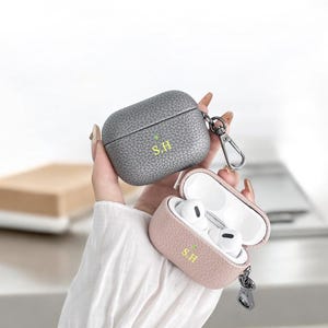 May include: Two leather Airpod cases with a keychain. The case on the left is gray and the case on the right is pink. Both cases have the initials "S.H." embossed on them.