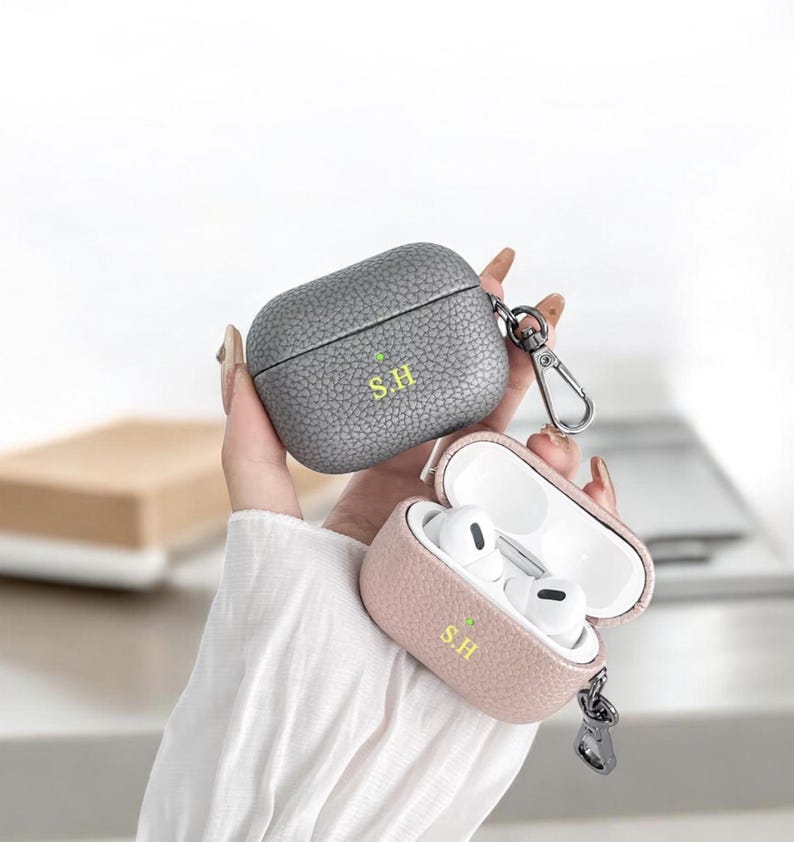 May include: Two leather Airpod cases with a keychain. The case on the left is gray and the case on the right is pink. Both cases have the initials "S.H." embossed on them.