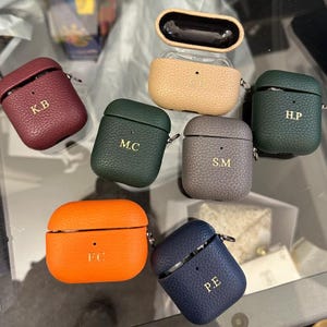 May include: Seven different colored leather Airpod cases with personalized initials. The cases are in shades of burgundy, green, orange, gray, blue, and tan.