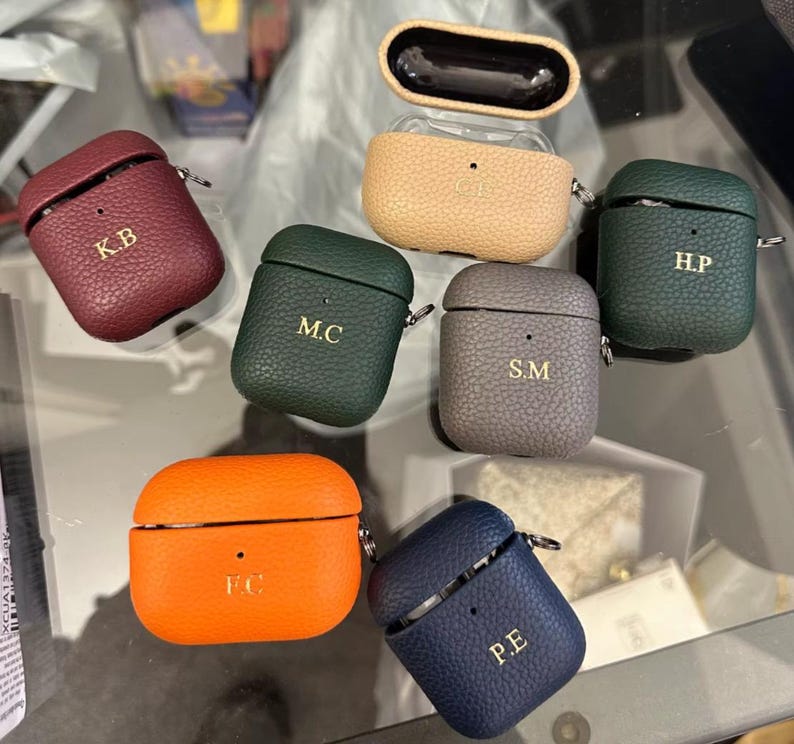 May include: Seven different colored leather Airpod cases with personalized initials. The cases are in shades of burgundy, green, orange, gray, blue, and tan.