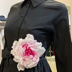 May include: A black dress shirt with a white collar and long sleeves. The shirt has a large pink and white flower attached to the waist.