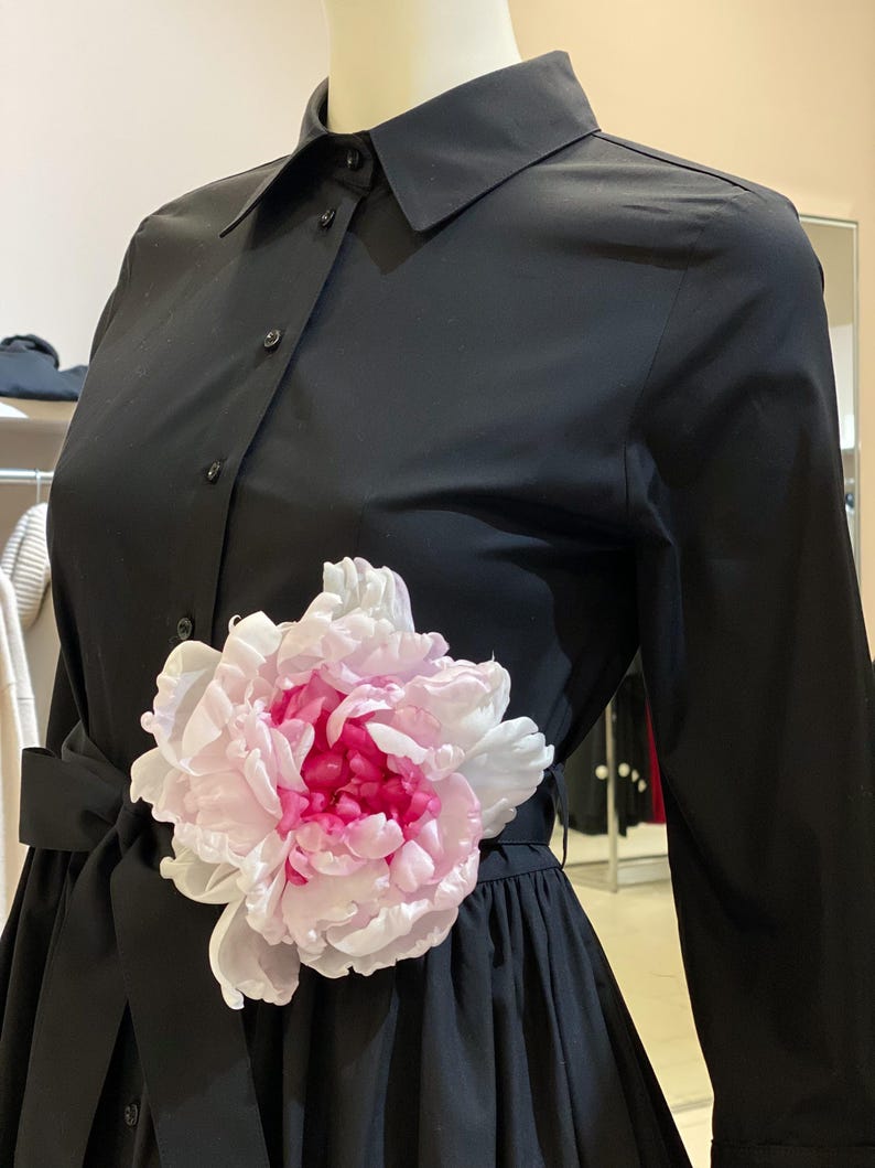 May include: A black dress shirt with a white collar and long sleeves. The shirt has a large pink and white flower attached to the waist.