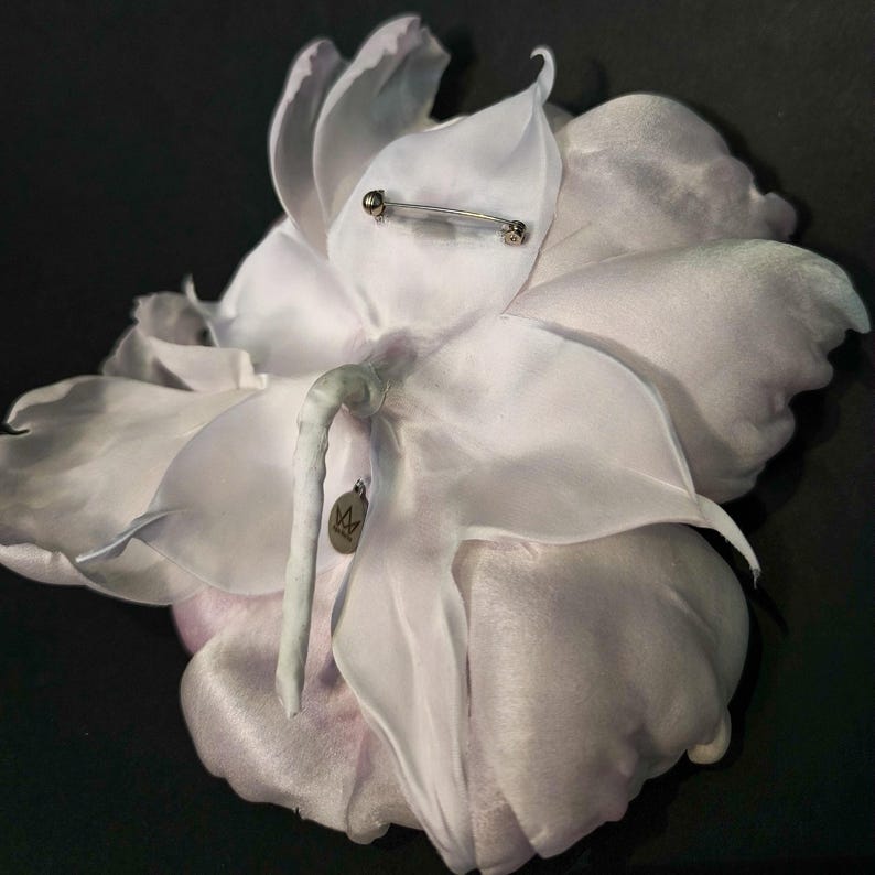 May include: A white silk flower brooch with a silver pin and a small tag with a crown logo. The flower has a delicate, layered design and a soft, feminine feel.