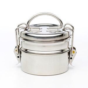 May include: A silver stainless steel lunch box with a handle and two tiers. The lunch box has a polished finish and is designed for carrying food.