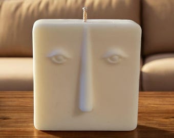 May include: A white square candle with a face sculpted into the front. The face has two eyes and a long nose.
