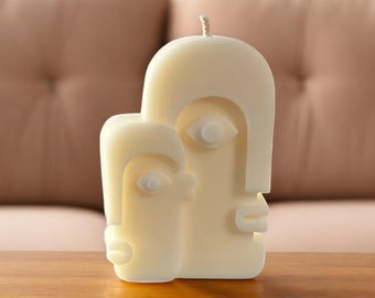 May include: A white candle shaped like two abstract faces, one behind the other. The candle has a simple, modern design with geometric features.