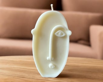 May include: White candle shaped like a face with a single eye and nose. The candle is on a wooden surface.