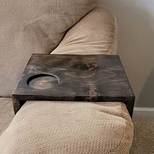 May include: A dark stained wooden sofa arm table with a circular cutout for a cup or glass. The table is rectangular and sits on the arm of a beige upholstered sofa. The wood grain is visible.