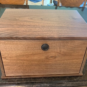 May include: A rectangular wooden bread box with a dark walnut finish. The box features a drawer with a round, black metal handle. The wood grain is visible on the front and top surfaces.