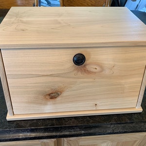 May include: A wooden bread box with a single door and a black knob. The box is made of light wood with a natural finish.