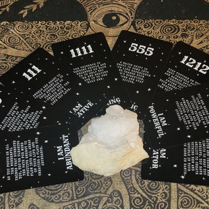 Puede incluir: Un juego de cartas negras con texto blanco y estrellas. Cada carta presenta una secuencia de números diferente y un mensaje. Las cartas están dispuestas en forma de abanico alrededor de un cristal blanco.