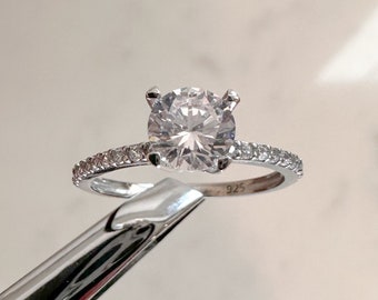 May include: A silver ring with a large, clear round cubic zirconia stone and a band of smaller cubic zirconia stones. The ring is marked "925" on the inside of the band.