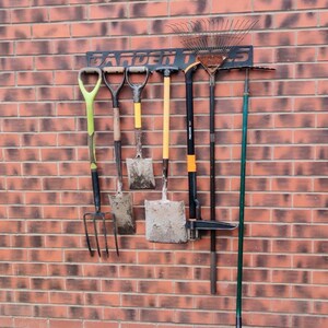 May include: A black metal garden tool rack with the words "GARDEN TOOLS" printed on it. The rack is mounted on a brick wall and holds a variety of tools, including a rake, a shovel, a hoe, and a pitchfork.